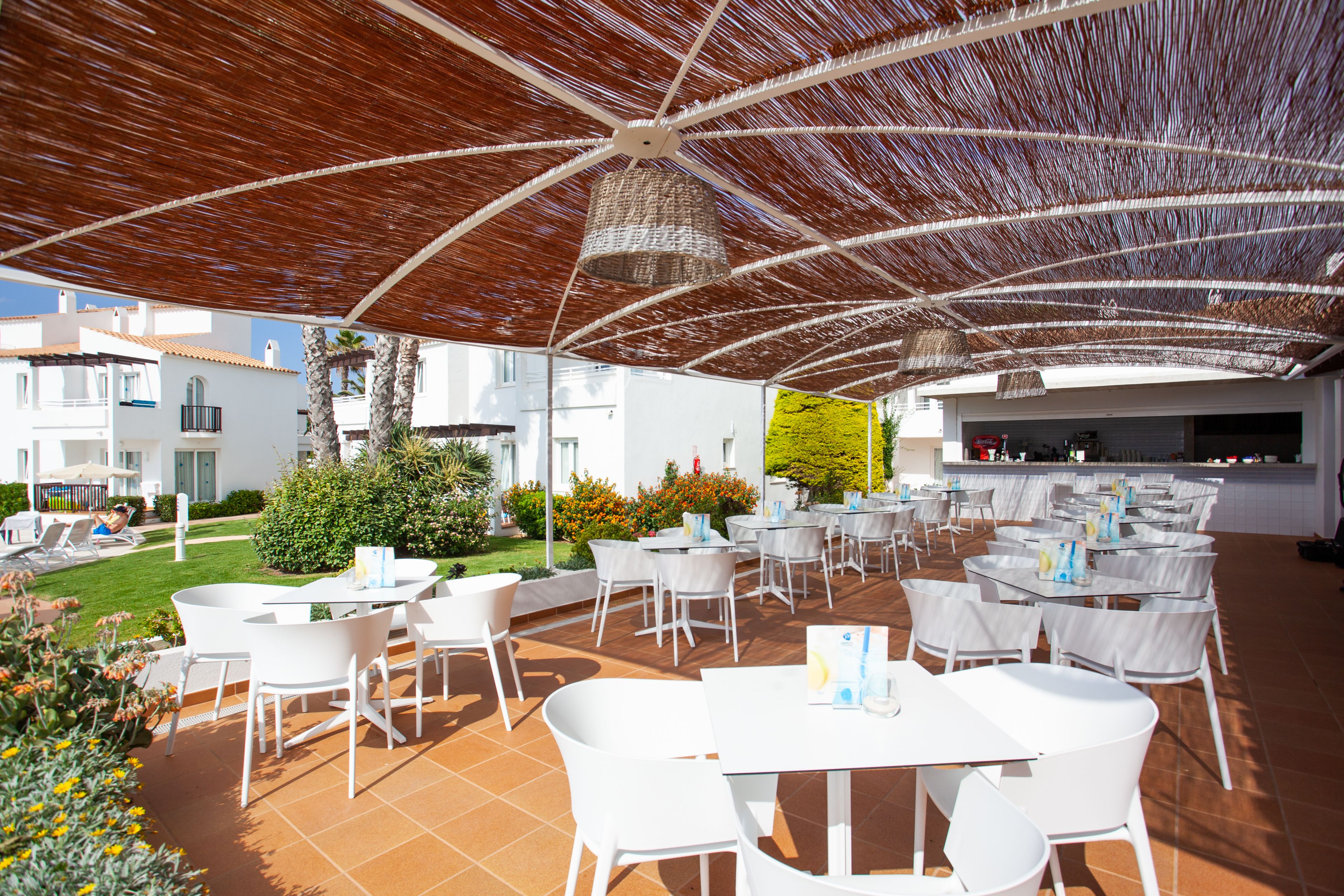 Un espacio de comedor al aire libre con mesas y sillas blancas, una pérgola en la parte superior y vistas de los edificios circundantes.