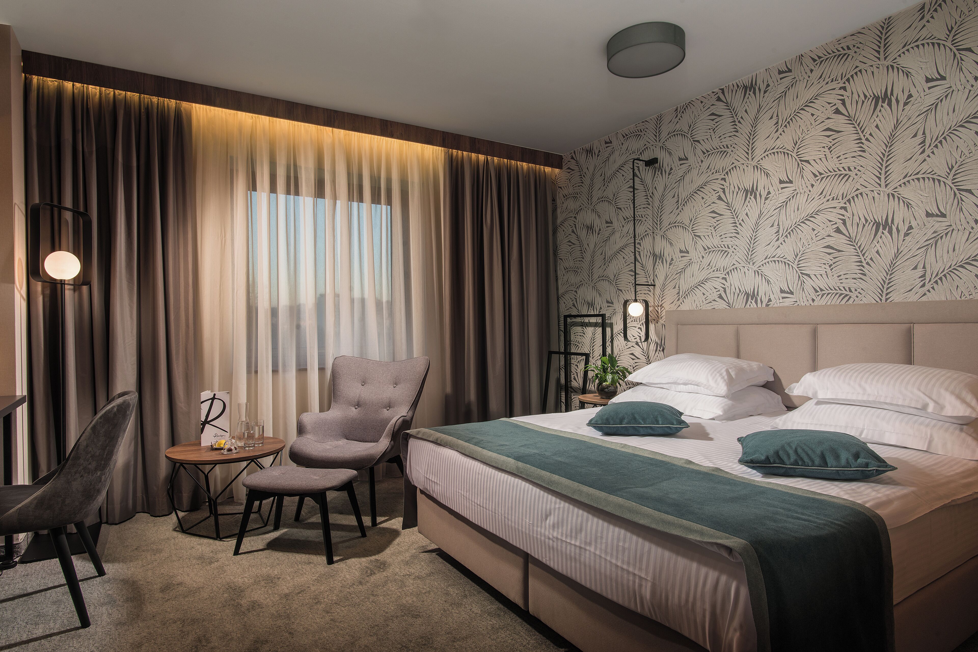 Habitación de hotel con cama, dos sillas, una mesita con lámpara y un ventanal con cortinas.