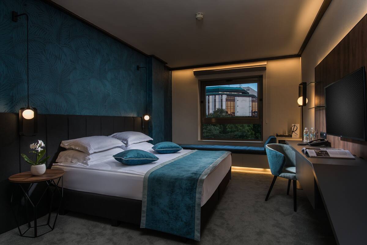 A modern bedroom with a bed, bedside table, chair, desk, and window.