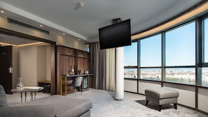 Habitación de hotel moderna con un ventanal grande, televisión de pantalla plana, un sofá, una pequeña mesa y una estantería.