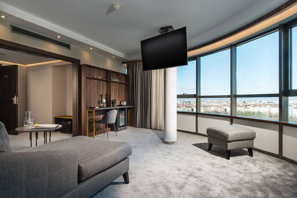A modern hotel room with a large window, a flat-screen TV, a sofa, a small table, and a bookshelf.