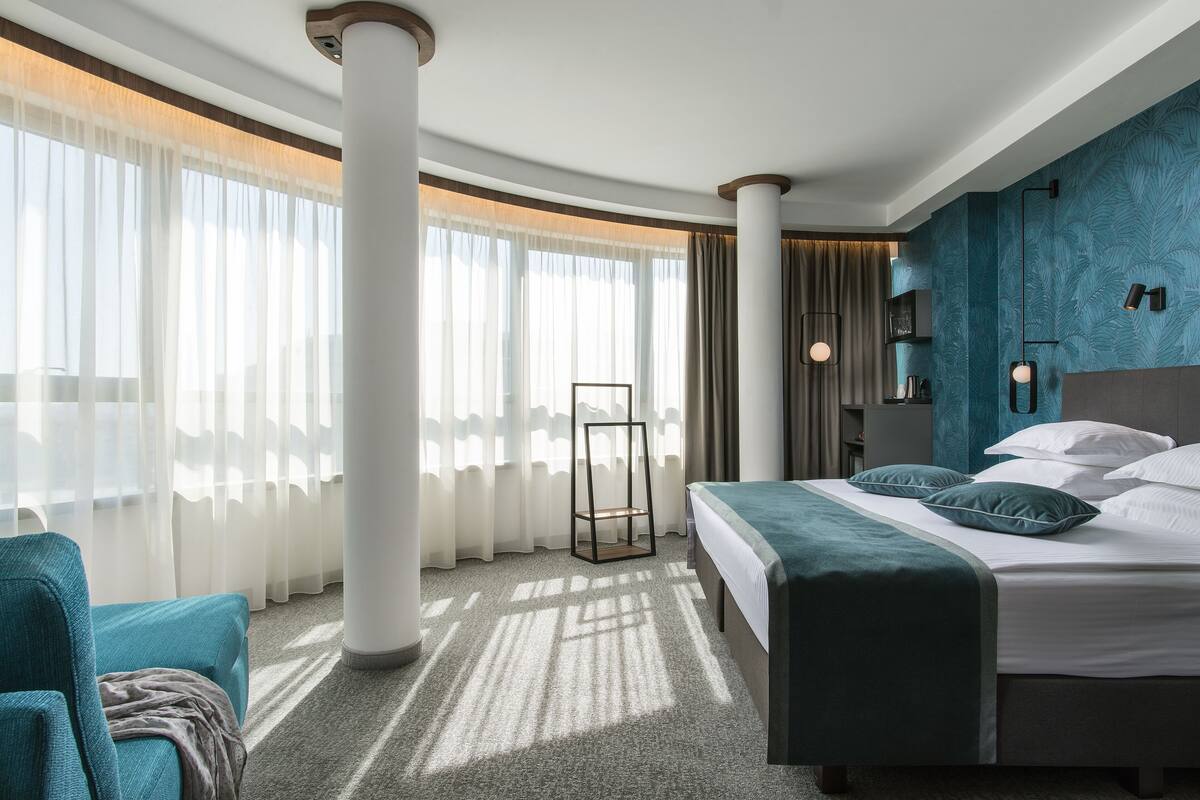 A hotel room with a bed, a chair, a standing lamp, and a large window with sheer curtains.