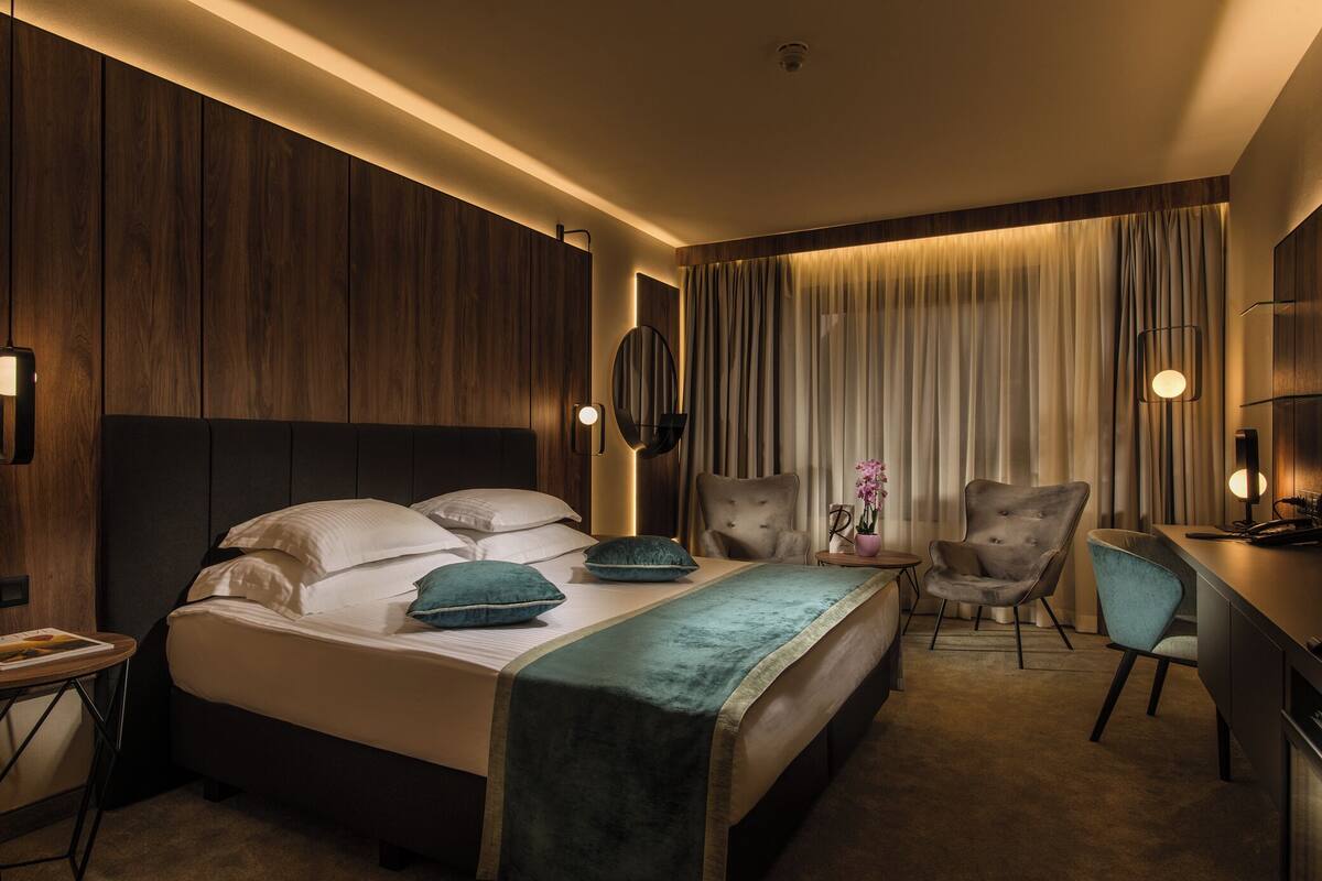 A hotel room with a bed, two chairs, a desk, and a large window with curtains.