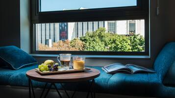 Una habitación con un ventanal amplio, una mesa redonda con una bandeja de frutas y un vaso de jugo de naranja, un libro y un sofá azul.