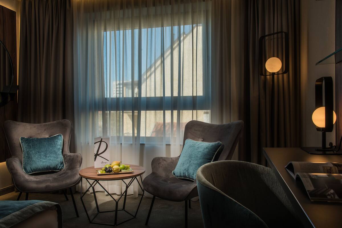 A modern hotel room with two armchairs, a small round table with fruit, and a large window with curtains.