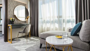 Una habitación de hotel moderna con un sofá gris, una mesita de centro redonda, un escritorio con una silla y un amplio ventanal con cortinas traslúcidas.
