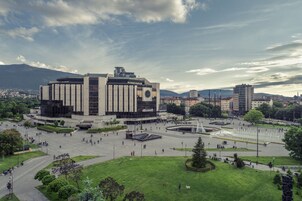A large public square with a central building, fountains, and surrounding greenery.