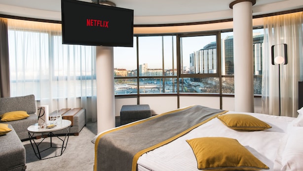 Habitación de hotel con una cama grande, un sofá, una mesita y un televisor en la pared mostrando 'NETFLIX'.