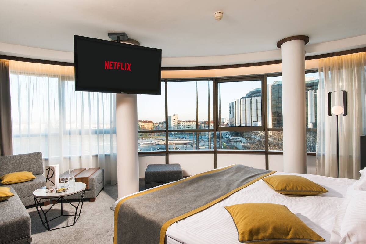 A hotel room with a large bed, a sofa, a small table, and a TV mounted on the wall displaying 'NETFLIX'.