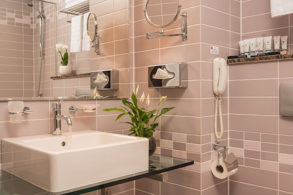 Un baño con un lavamanos blanco, un estante de vidrio, una planta en maceta, un perchero, un dispensador de papel higiénico y un teléfono.