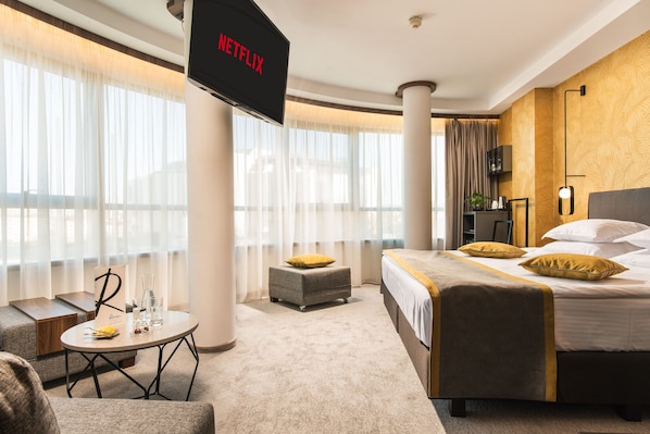A modern hotel room with a bed, a small table with a sign, a TV displaying 'NETFLIX', and large windows with sheer curtains. - Rosslyn Central Park Hotel (Sofia)