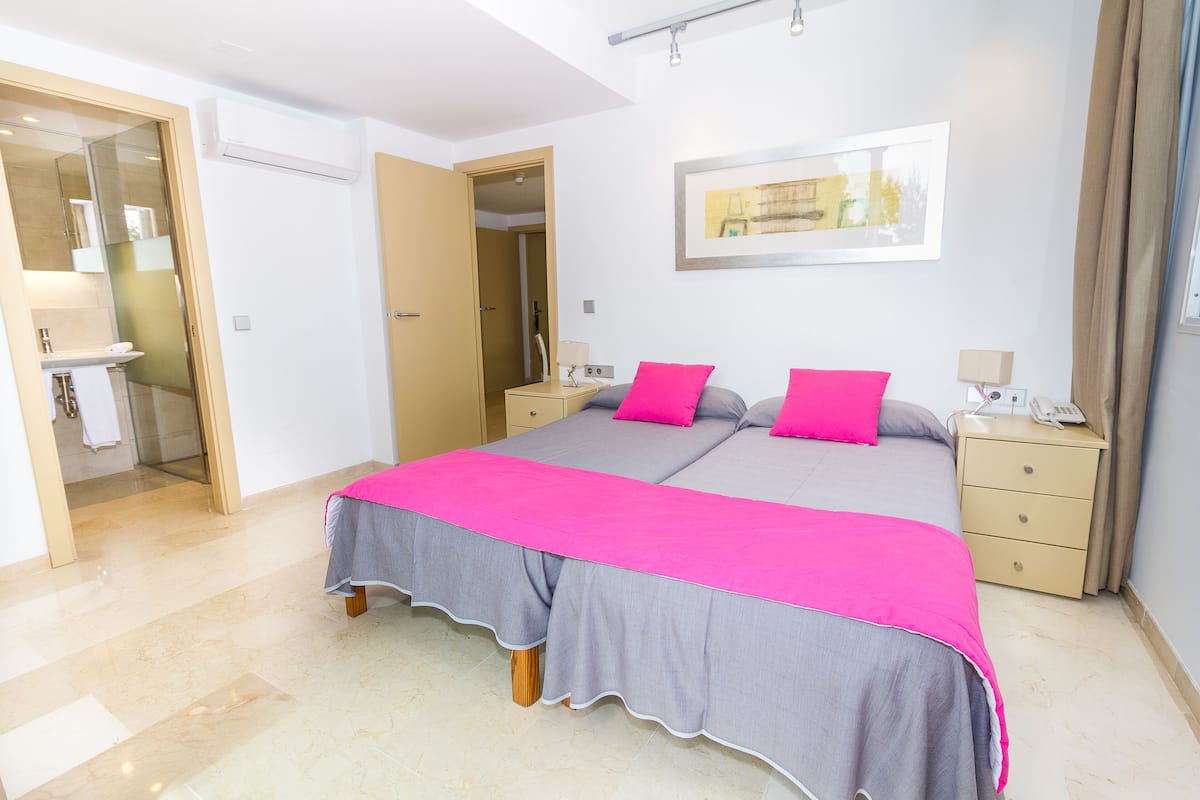 Una cama doble con colcha gris y rosa, una mesita de noche con lámpara y un cuadro colgado en la pared.