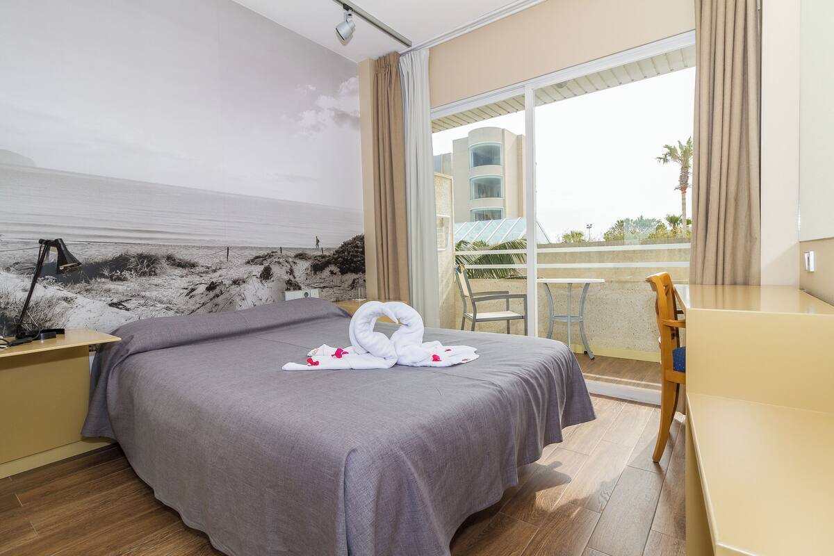 Habitación de hotel con cama, escritorio, silla y balcón con vista a la playa y palmeras.