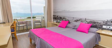 Un dormitorio con una cama grande, un escritorio y una silla. Se ve la playa a través de un ventanal amplio.