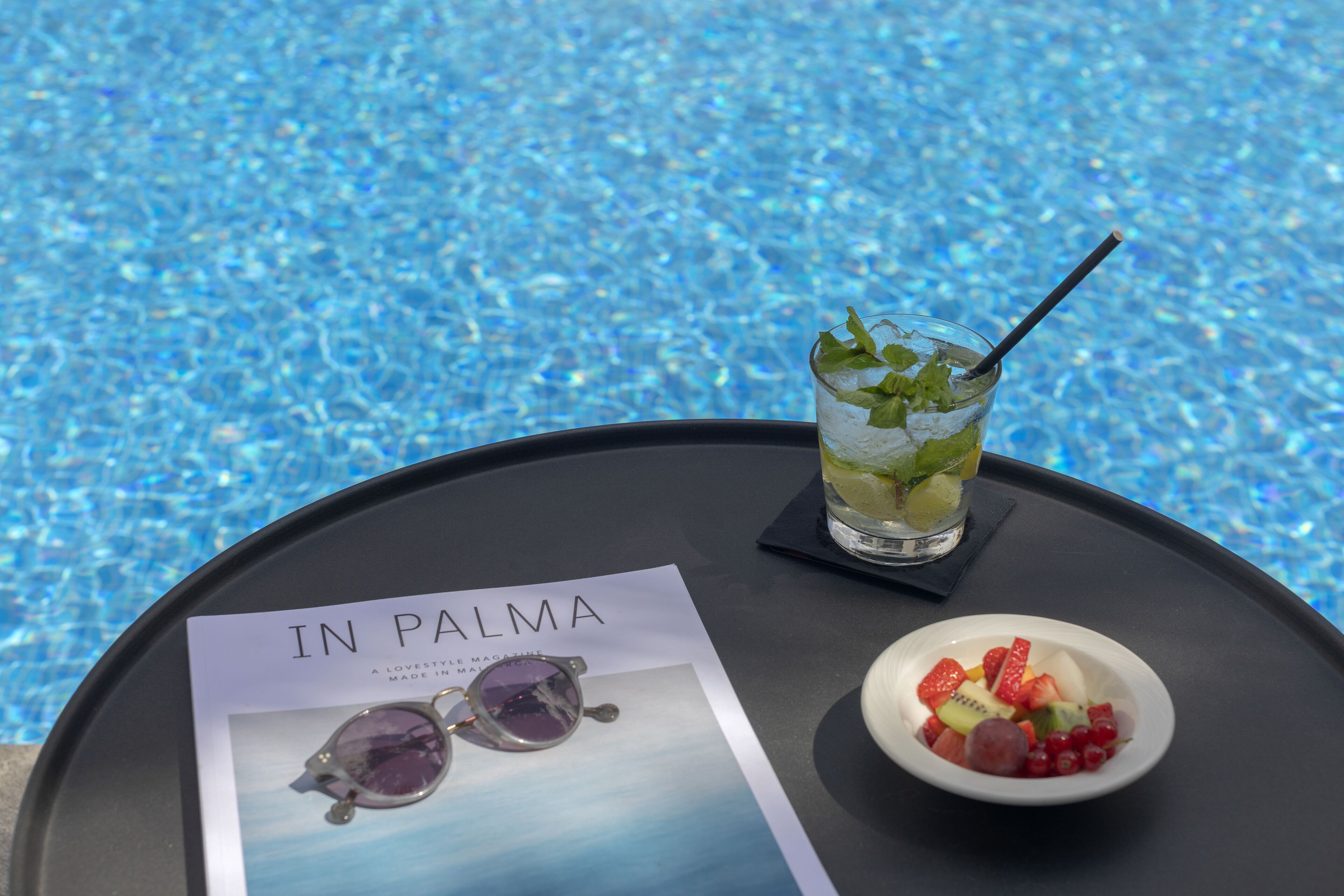Bandeja con una bebida, un cuenco de fruta y una revista cerca de una piscina.