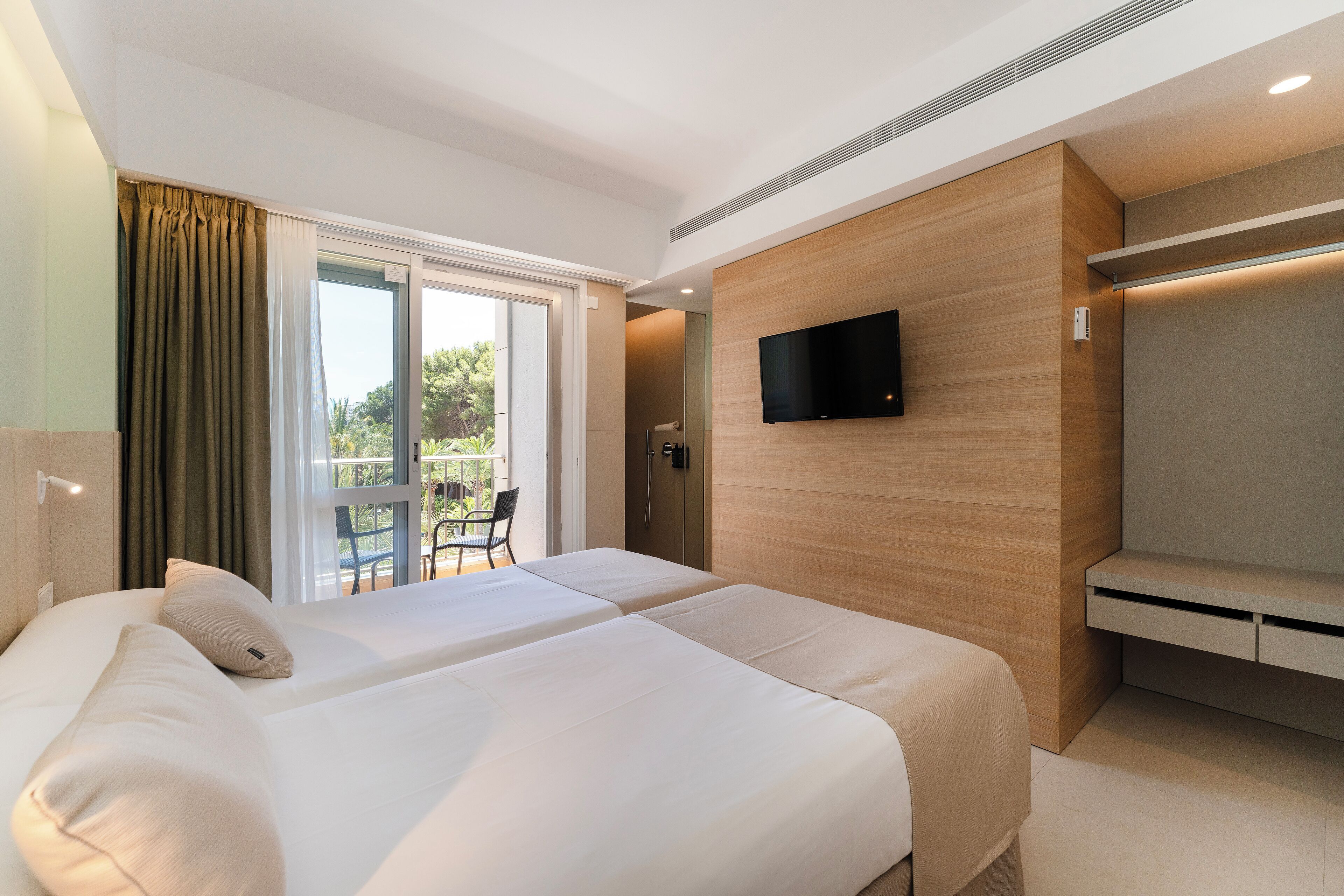 Een hotelkamer met twee bedden, een aan de muur bevestigde televisie en een schuifdeur die leidt naar een buitenruimte met een tafel en stoelen.