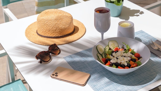 Una mesa con ensalada, un sombrero, gafas de sol, un teléfono y una bebida.