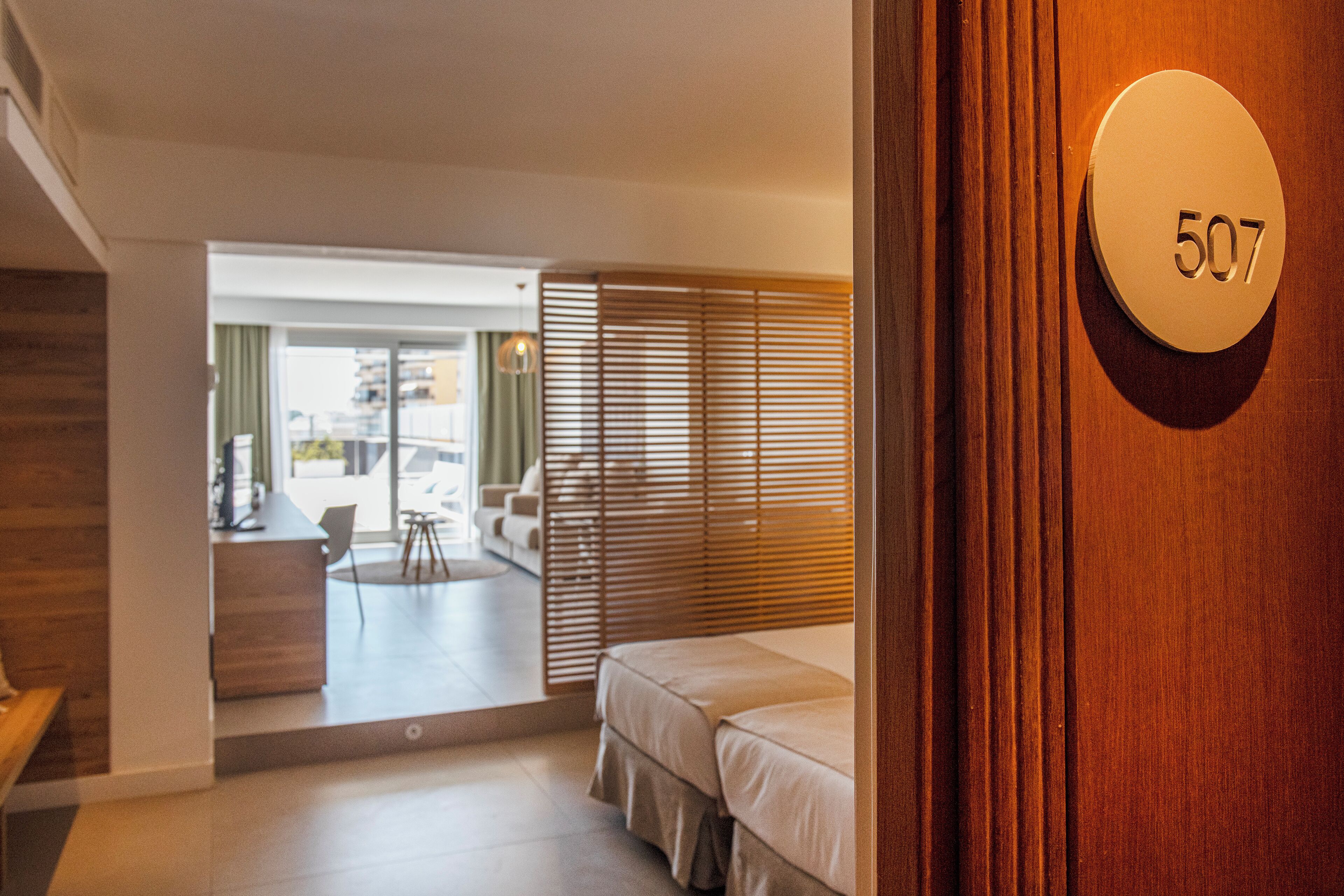 A hotel room with a wooden door, a number plate '507', a bed, a desk, and a view of the outside.
