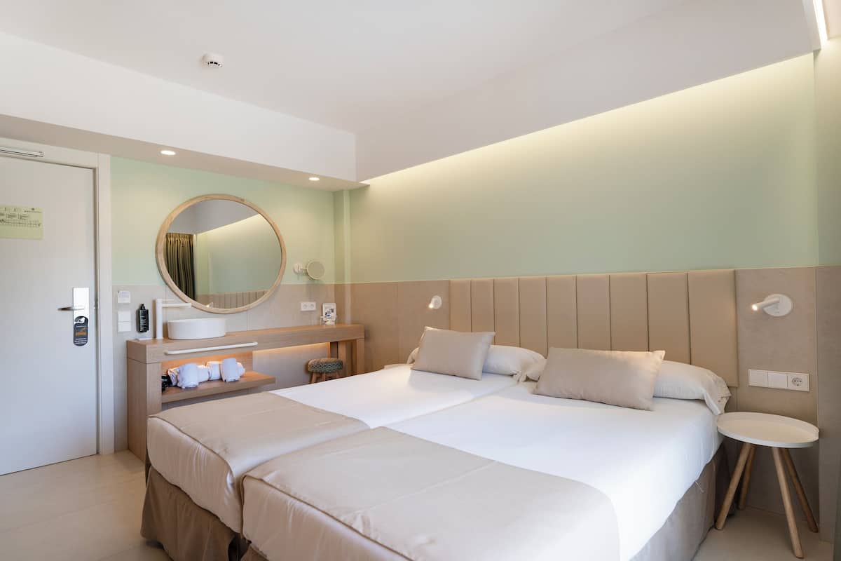 A hotel room with a large bed, a round mirror, a bedside table, and a small stool.
