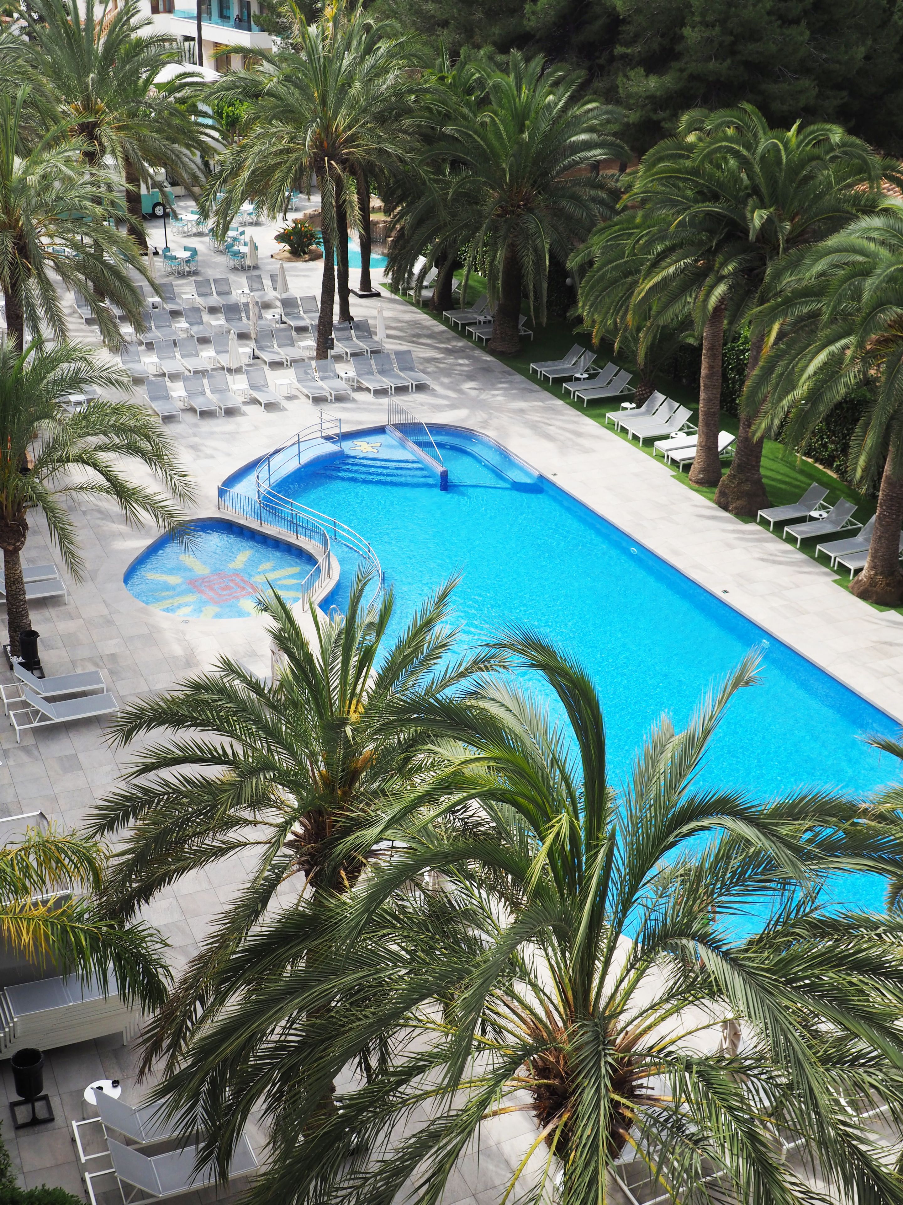 A large rectangular swimming pool with a blue center, surrounded by lounge chairs and palm trees.
