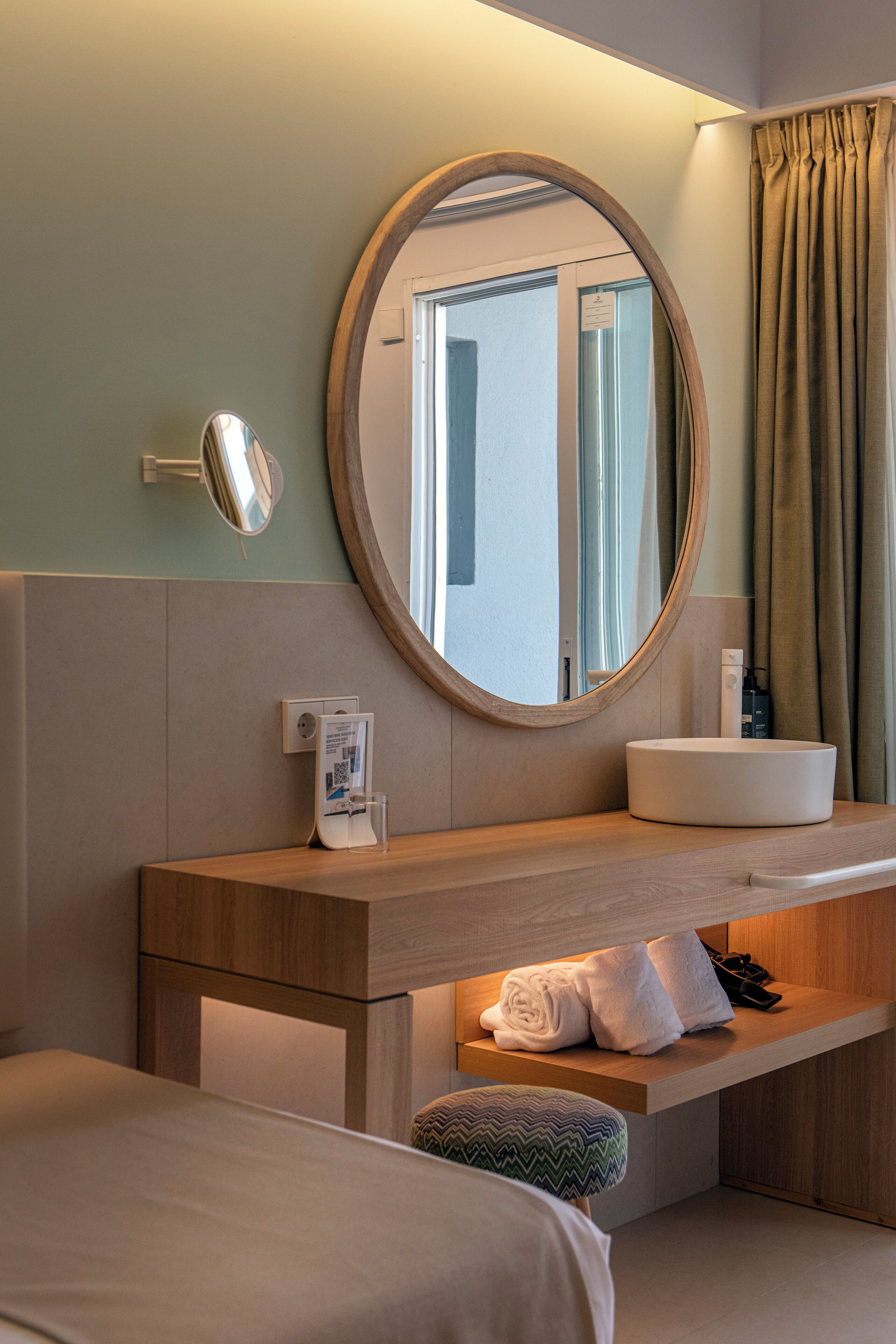 A hotel room with a wooden vanity, a round mirror, a sink, and a bed.