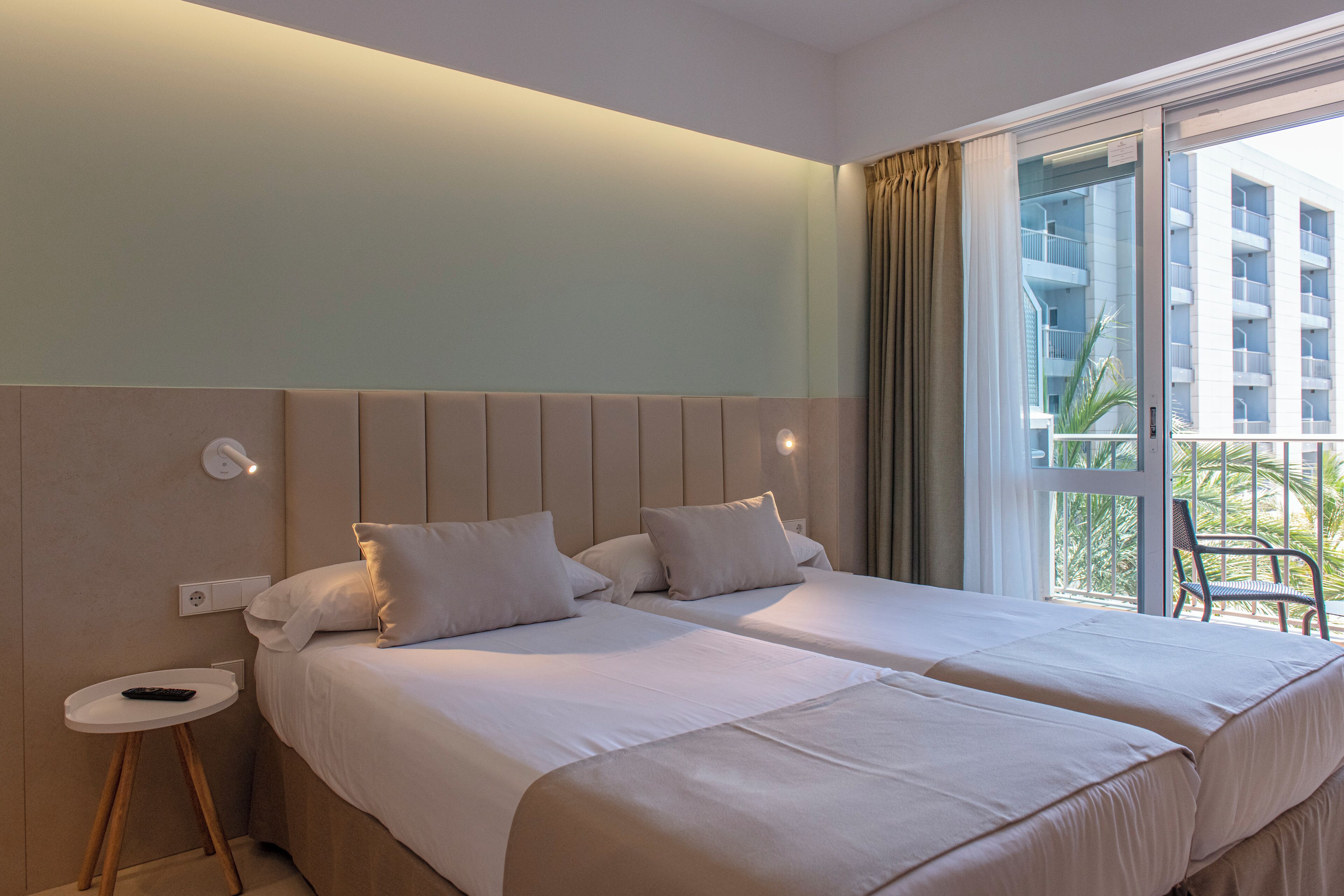 Habitación de hotel con dos camas, una mesita de noche, un enchufe y vistas a un edificio y palmeras a través de un ventanal.