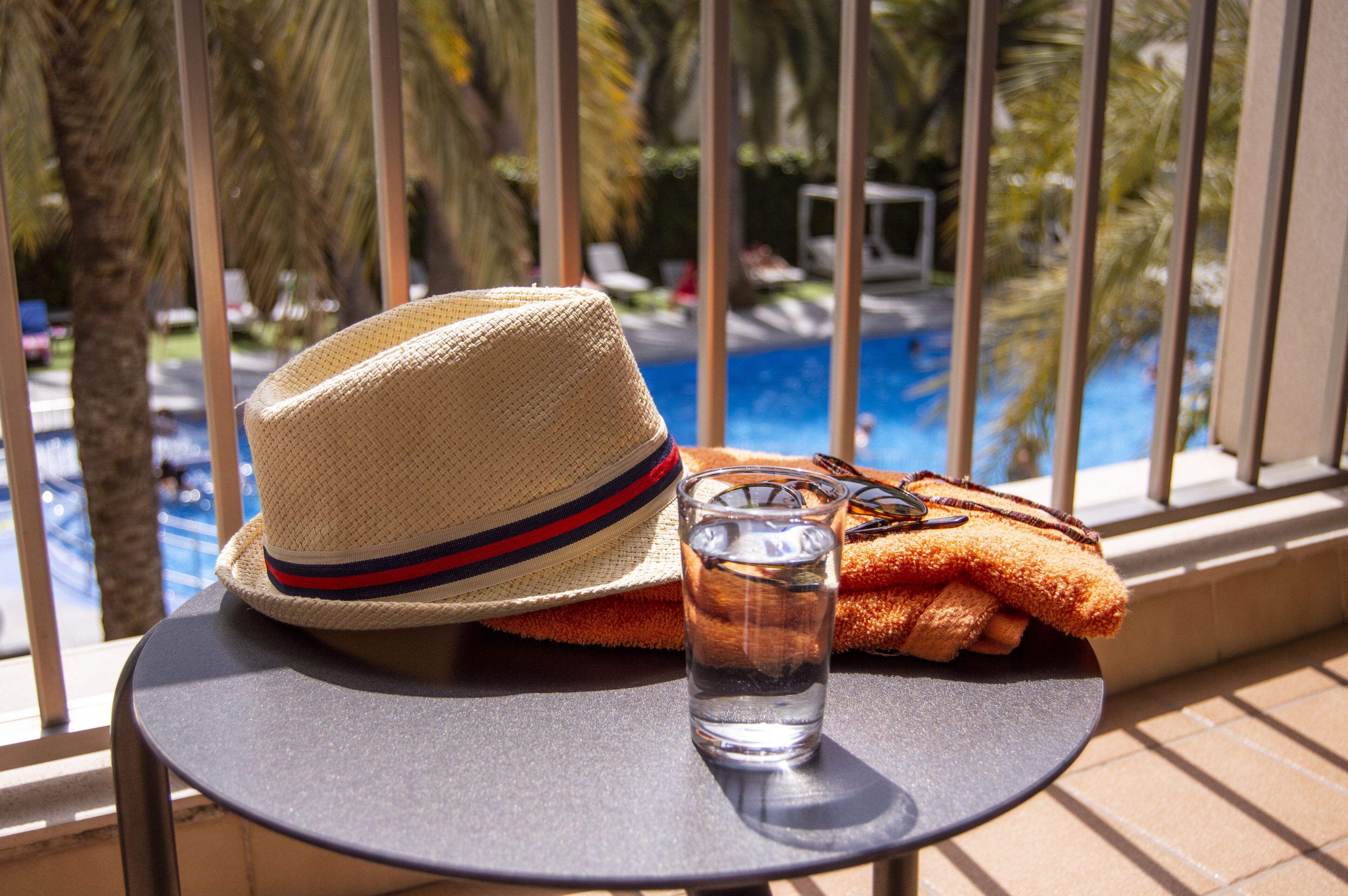 Ein Strohhut, ein Handtuch und ein Glas Wasser auf einem Balkon mit Blick auf ein Schwimmbad.