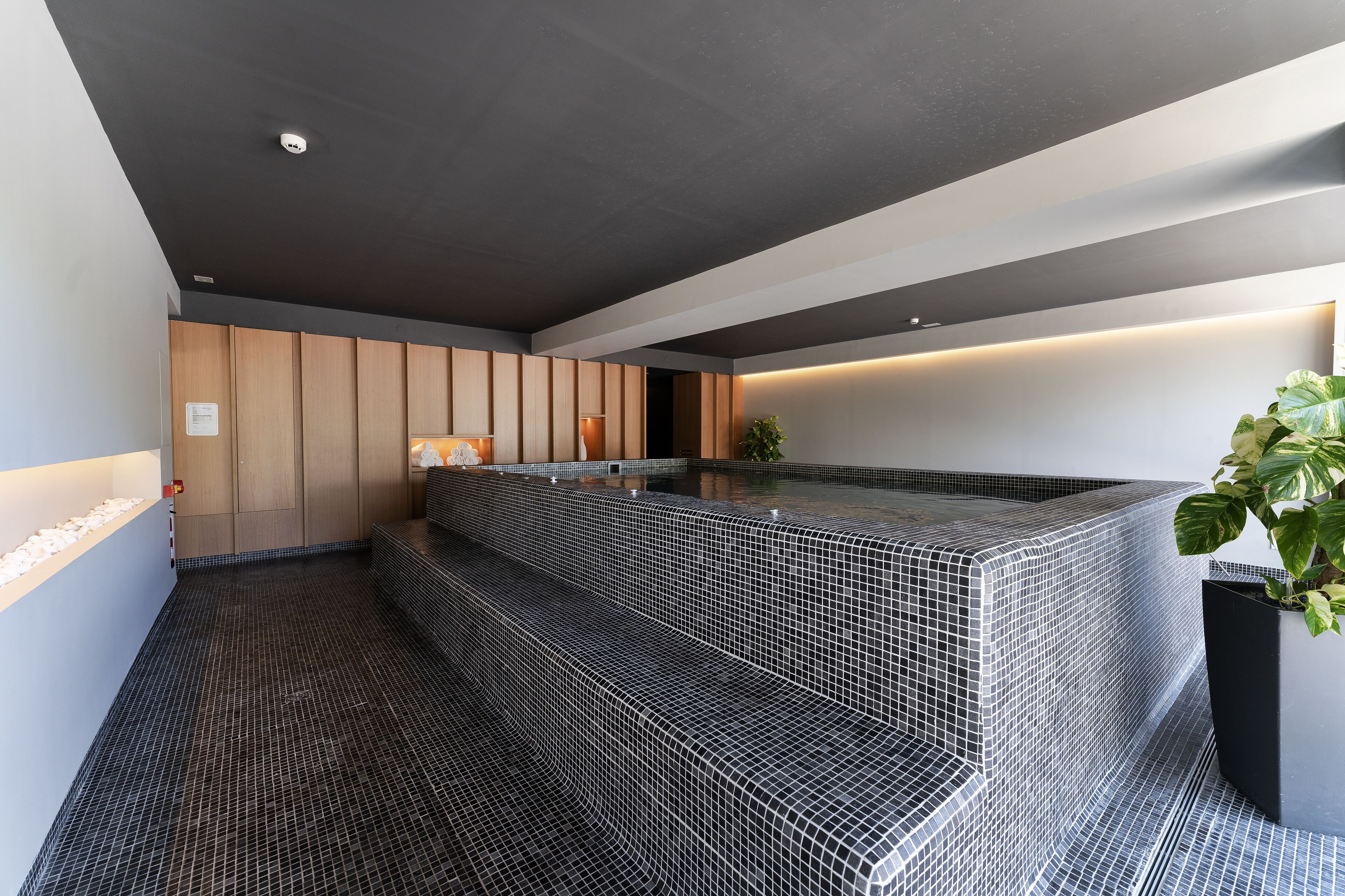 Un área de spa moderna con suelo de baldosas blanco y negro, una zona de descanso con banco y una planta en maceta.