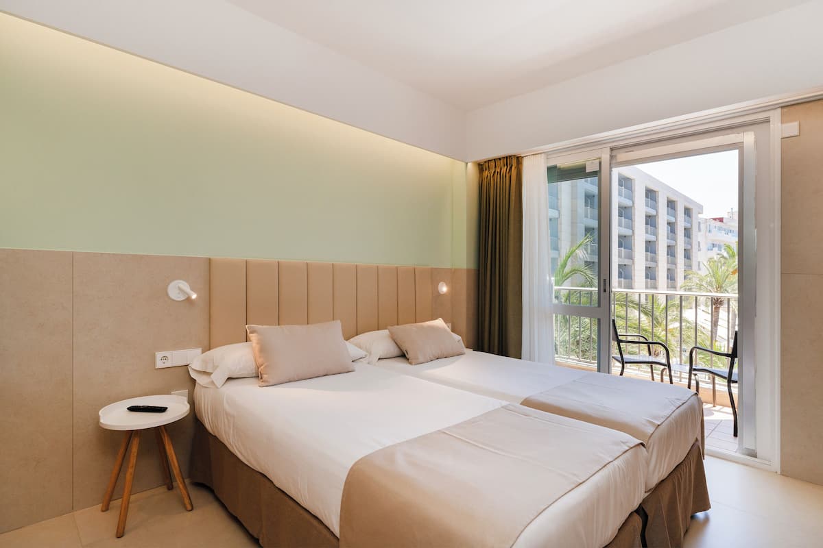 A hotel room with two beds, a small side table, a power outlet, and a balcony with a view of buildings and palm trees.