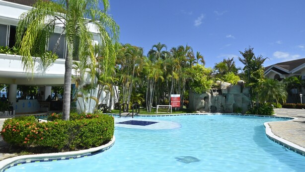 A swimming pool with a surrounding garden, palm trees, and a building with a white facade and a brown roof. - Hotel Palma Real (Jutiapa)