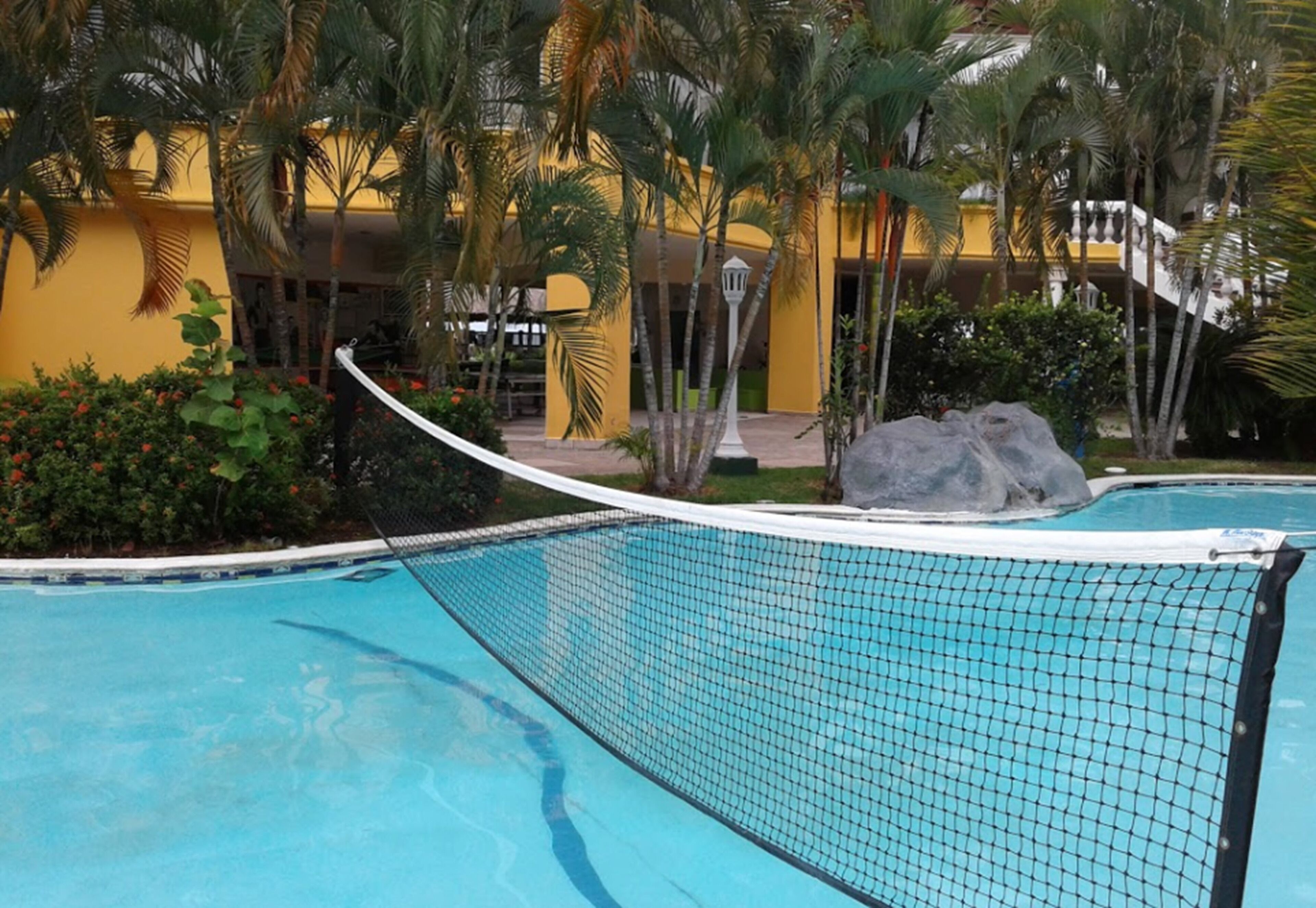 A swimming pool with a netted bridge, surrounded by palm trees and a yellow building.