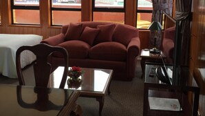 A hotel room with a wooden floor, a red sofa, a glass coffee table with a vase, a TV on a stand, and large windows with a city view. - Apart Hotel Colon (Puerto Montt)