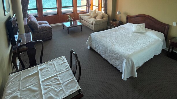 A hotel room with a bed, a sofa, a desk with a chair, and a view of the sea through large windows. - Apart Hotel Colon (Puerto Montt)