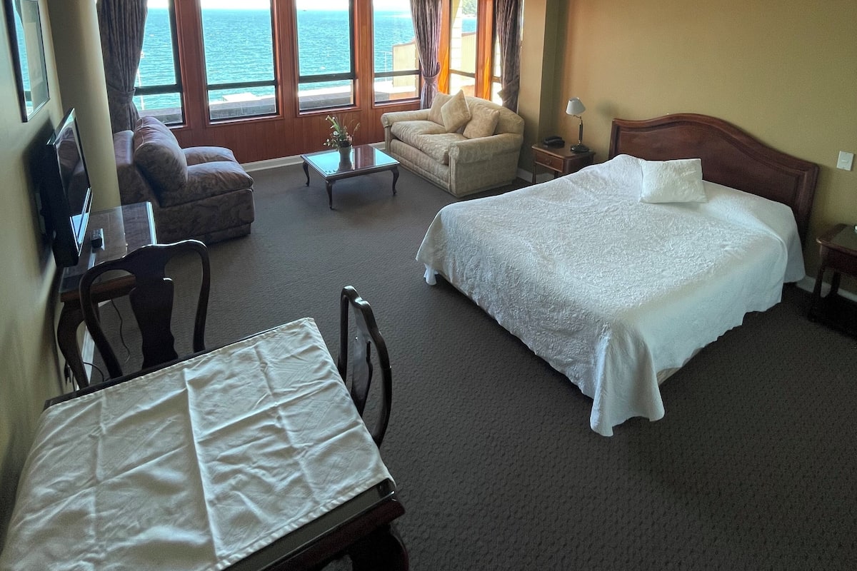 A hotel room with a bed, a sofa, a desk with a chair, and a view of the sea through large windows.