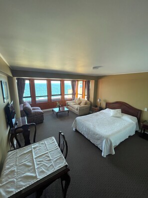 Habitación de hotel con cama, sofá, escritorio y silla, y vista al mar a través de amplios ventanales.