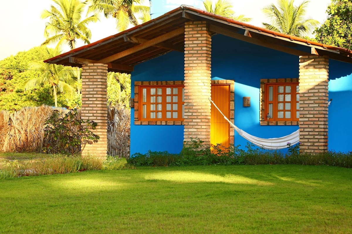 A small, single-story building with a blue exterior and brick pillars, featuring two windows with wooden shutters and a door.