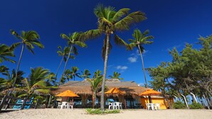 Un resort frente a la playa con cabañas de techo de paja, sillas de plástico blancas y palmeras.