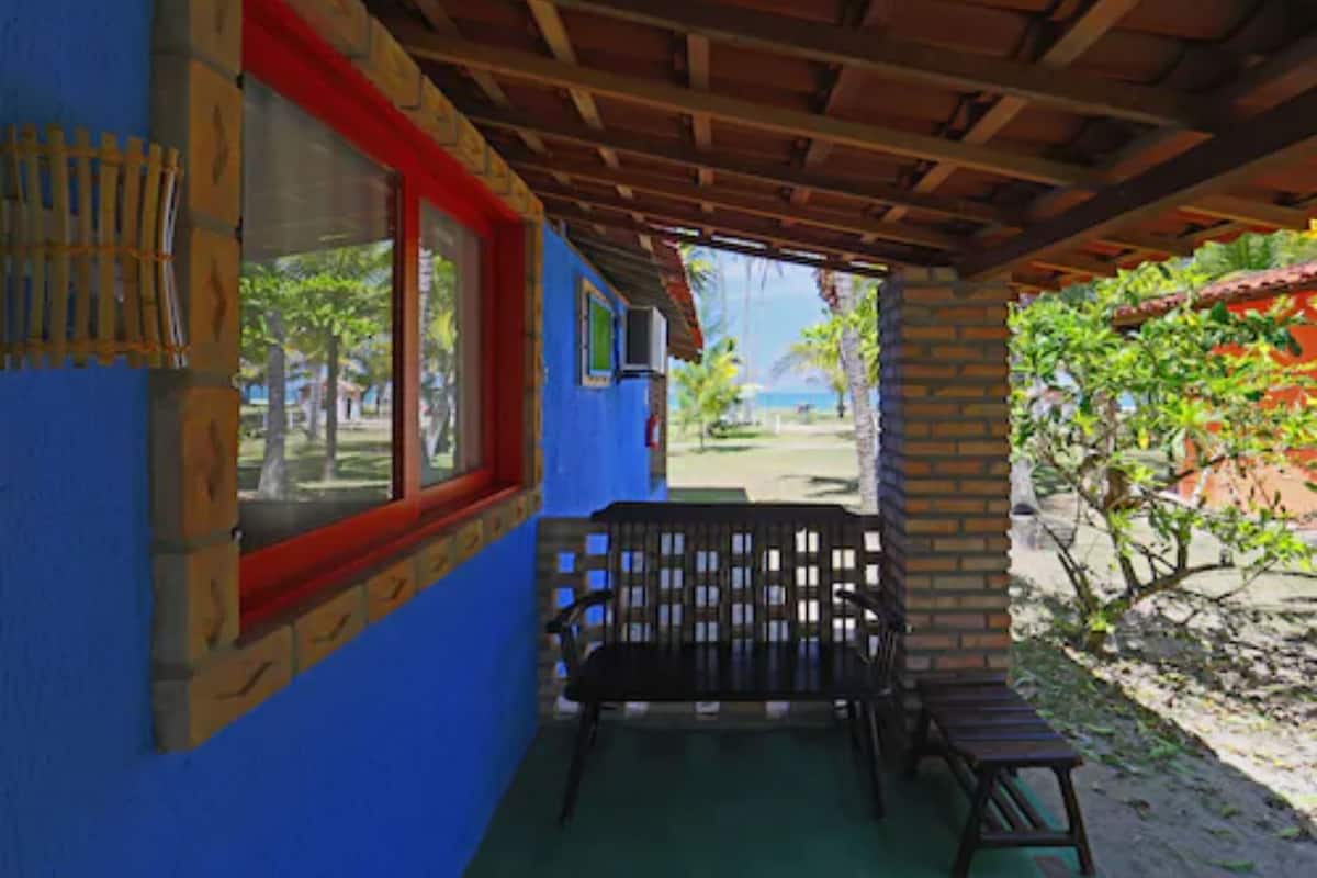 A covered outdoor seating area with a wooden bench, a table, and a view of the beach and ocean.