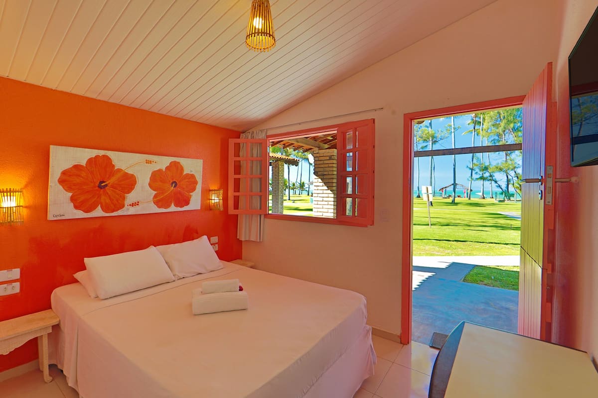 A hotel room with a large bed, a painting of flowers on the wall, and a view of a lawn and palm trees through an open door.