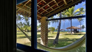 Vista a través de una ventana con una hamaca, palmeras y una construcción de techo de paja.