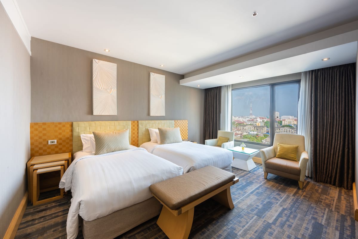 A hotel room with two beds, a bench, a small table, a chair, and a large window offering a city view.