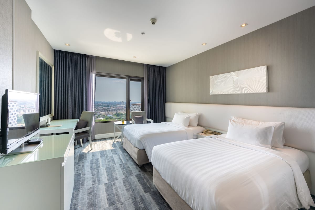 A hotel room with two beds, a desk, a chair, a TV, and a large window offering a city view.