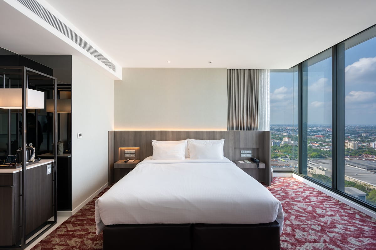 A hotel room with a large bed, a desk, a mirror, and a view of the city through large windows.