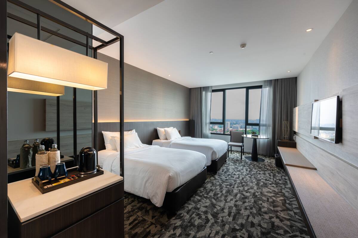 A hotel room with two beds, a desk, a chair, a large window, and a TV.