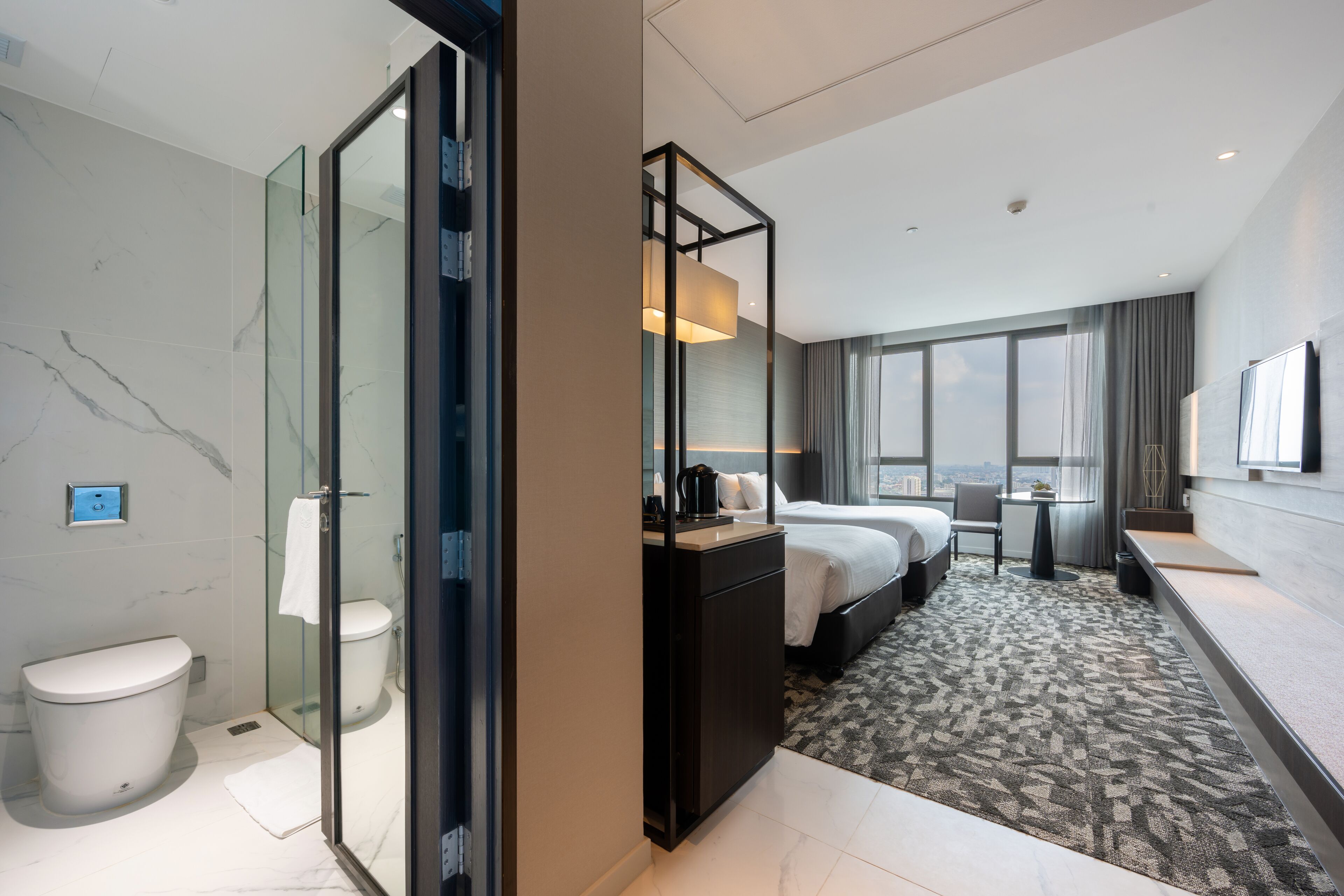 A modern hotel room with a glass shower enclosure, a bed with a headboard, a bedside table, a large window with a view, and a flat-screen TV mounted on the wall.
