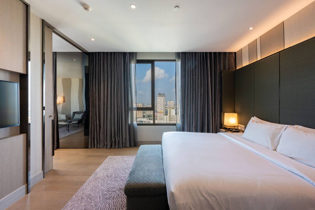 A modern hotel room with a large bed, a desk, and a view of the city through a large window.