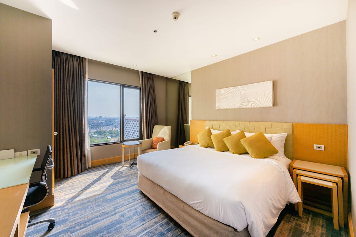 A hotel room with a large bed, a desk, a chair, a nightstand, and a view of the city.