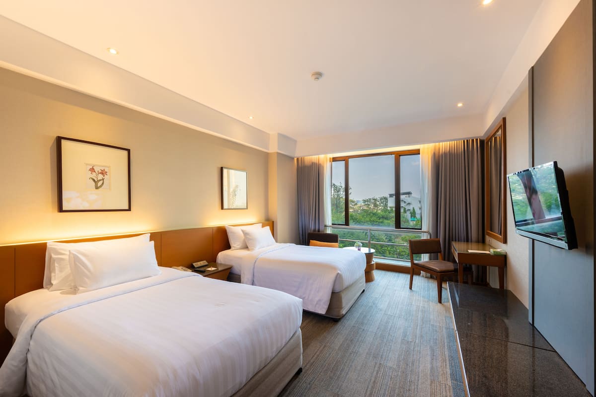 A hotel room with two beds, a desk, a chair, a TV, and a window with a view of greenery.