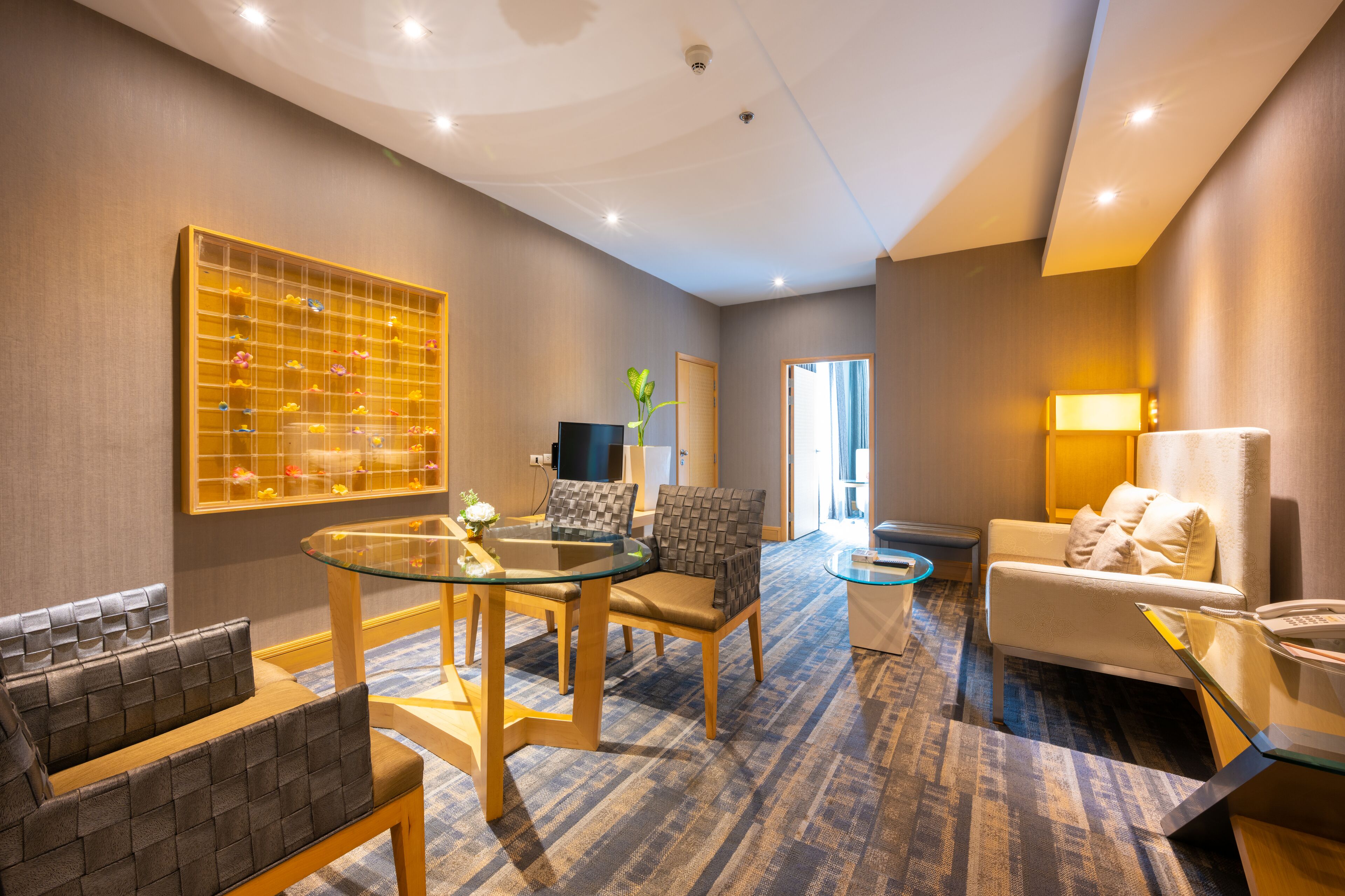 Una habitación de hotel moderna con una mesa de comedor de vidrio, asientos acolchados, un sofá y un televisor.