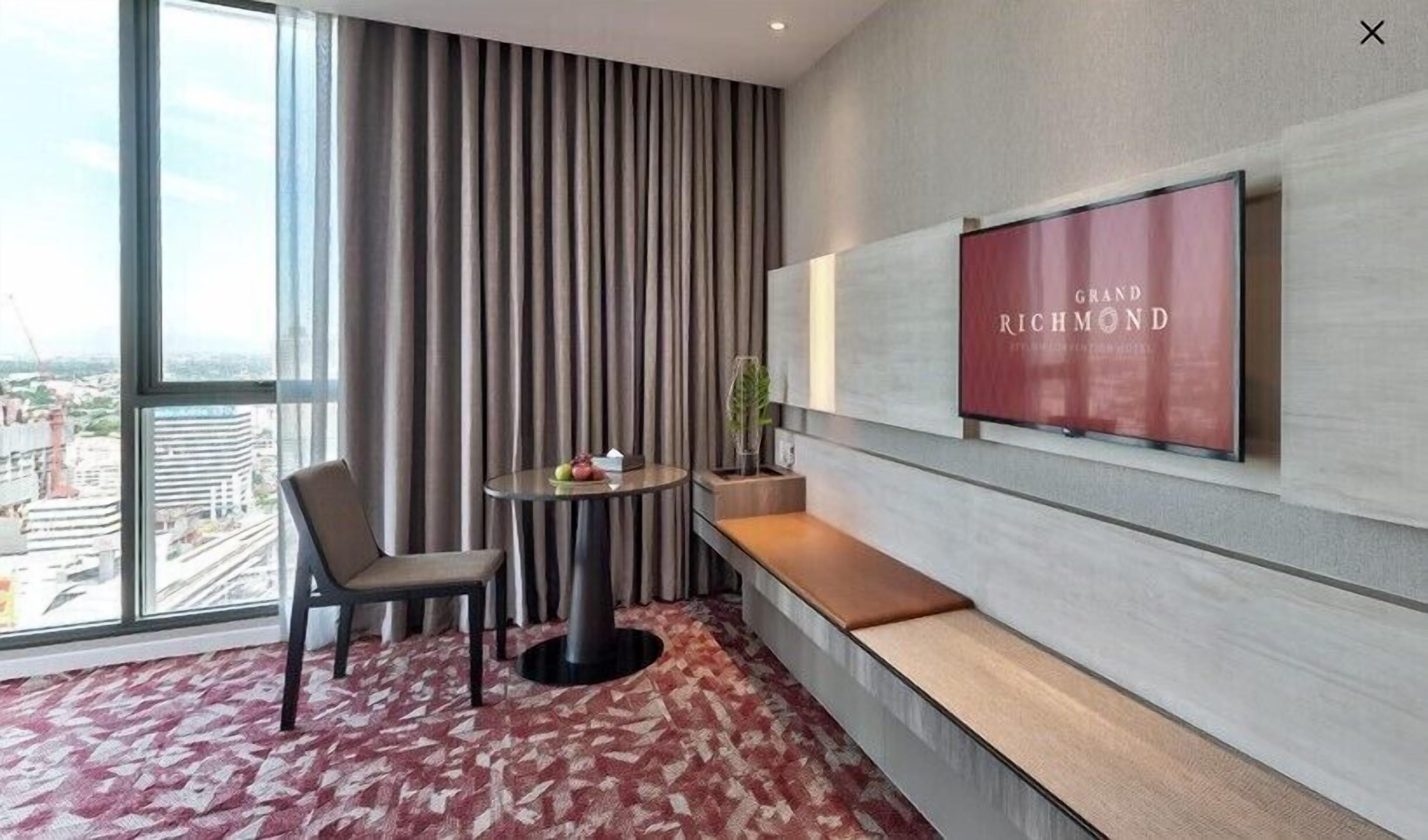 A hotel room with a large window, a TV mounted on the wall, a round table with a chair, and a bench.
